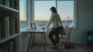 A person standing up from their desk beside an open laptop in a warm room at winter dusk, illustrating the transition from rest to action in ADHD task initiation.